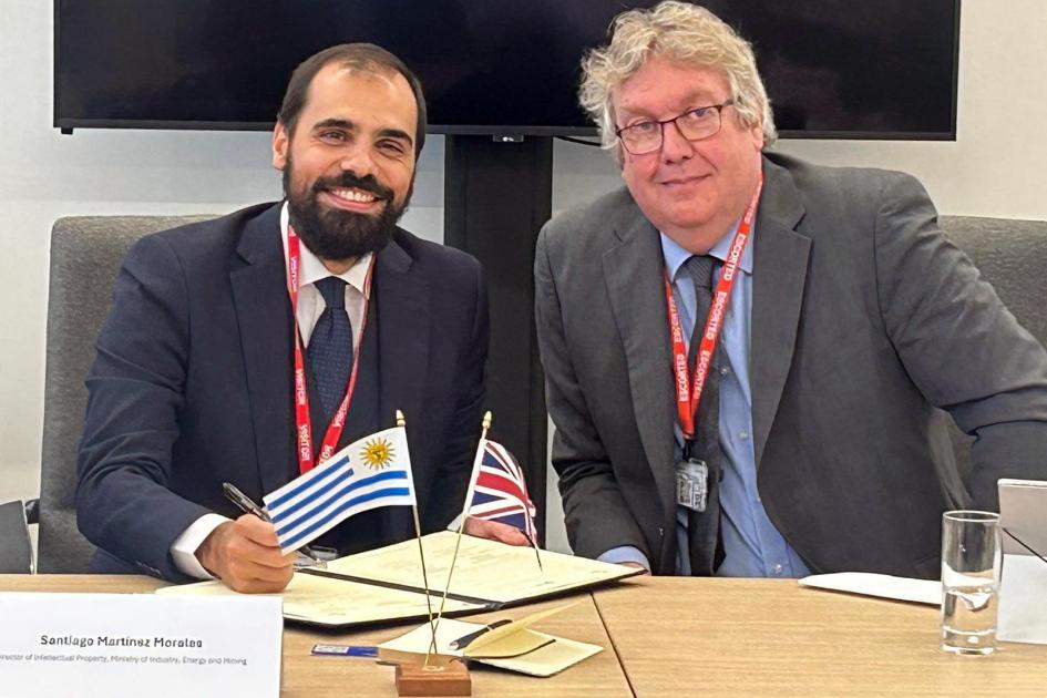 Santiago Martínez sonríe junto a un hombre; en la mesa hay banderas de Uruguay y el Reino Unidos