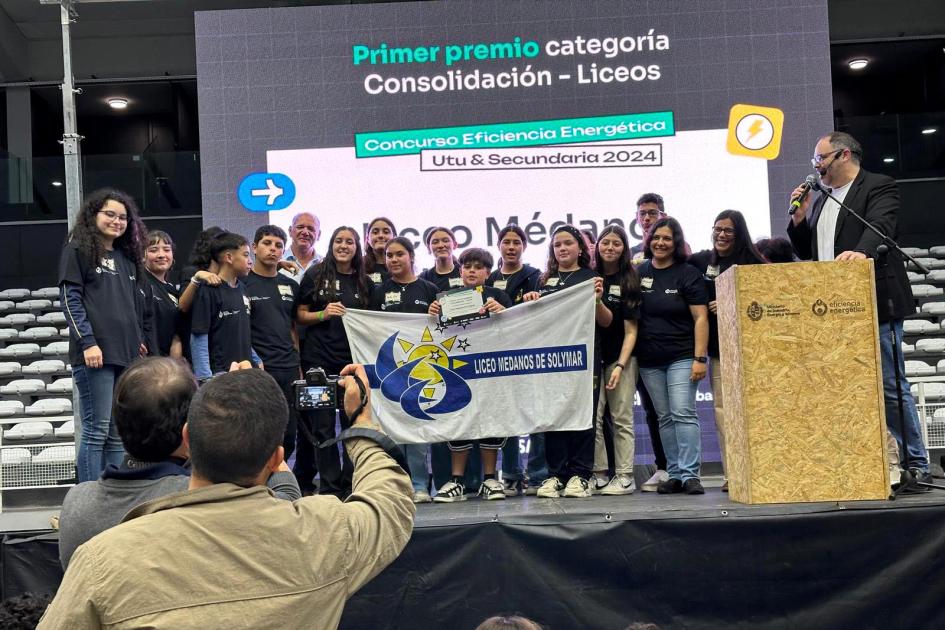 Ganadores de uno de los premios entregados Grupo sobre el escenario, con una bandera; detrás, pantalla con el nombre del premio