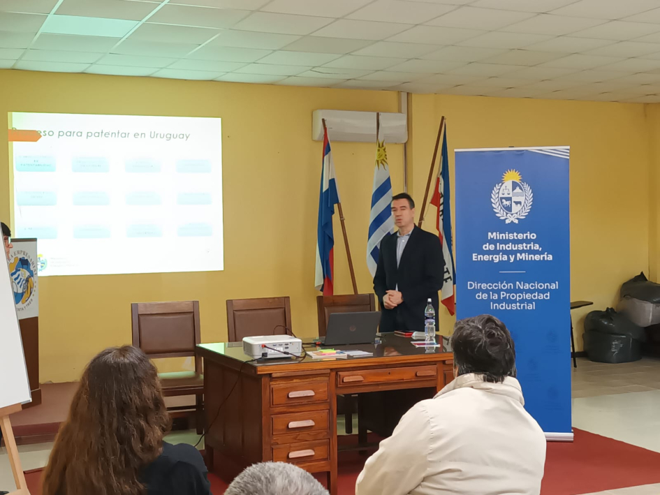 Presentación de Gabriel Wallusche Personas sentadas miran a un profesor; al fondo, pantalla, banner y banderas