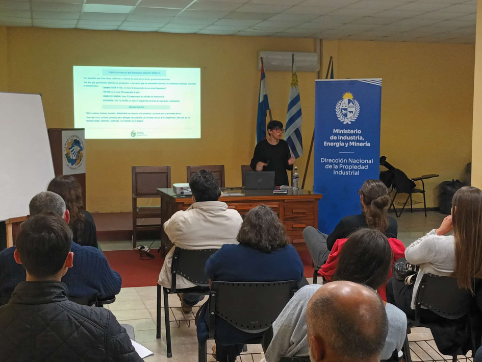 Presentación de Verónica Giménez Personas sentadas miran a un profesor; al fondo, pantalla, banner y banderas