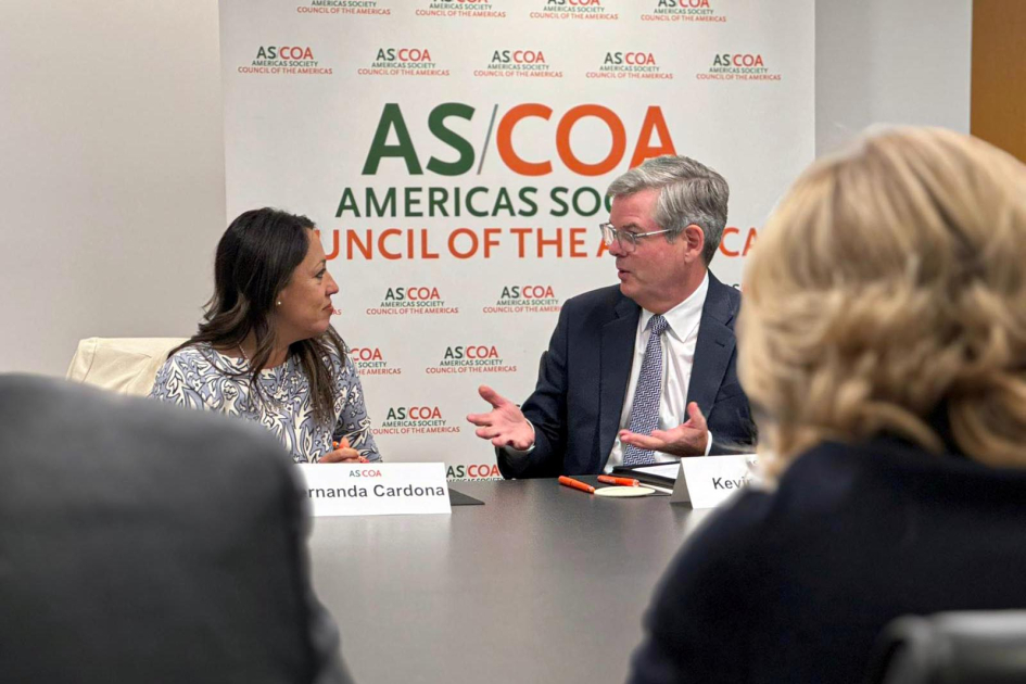 Reunión en AS/COA Ministra Cardona dialoga con un hombre; detrás, banner de AS/COA