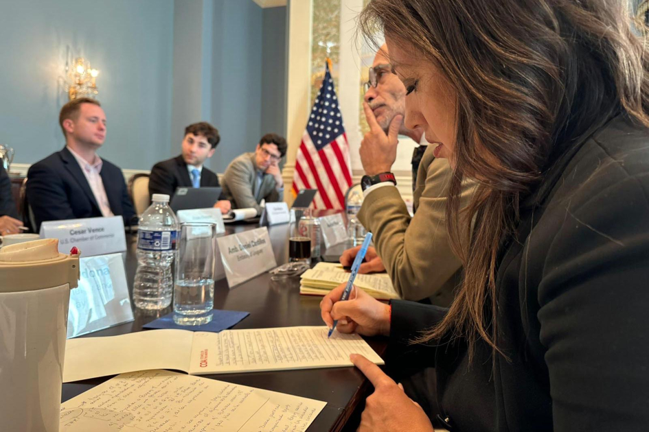 Reunión en Cámara de Comercio de Estados Unidos Cardona escribe en una mesa, junto a otras personas; al fondo, bandera de EE. UU.