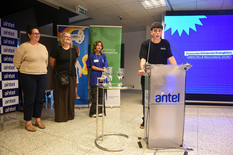 Estudiante haba a micrófono desde atril. Al fondo hay tres mujeres.