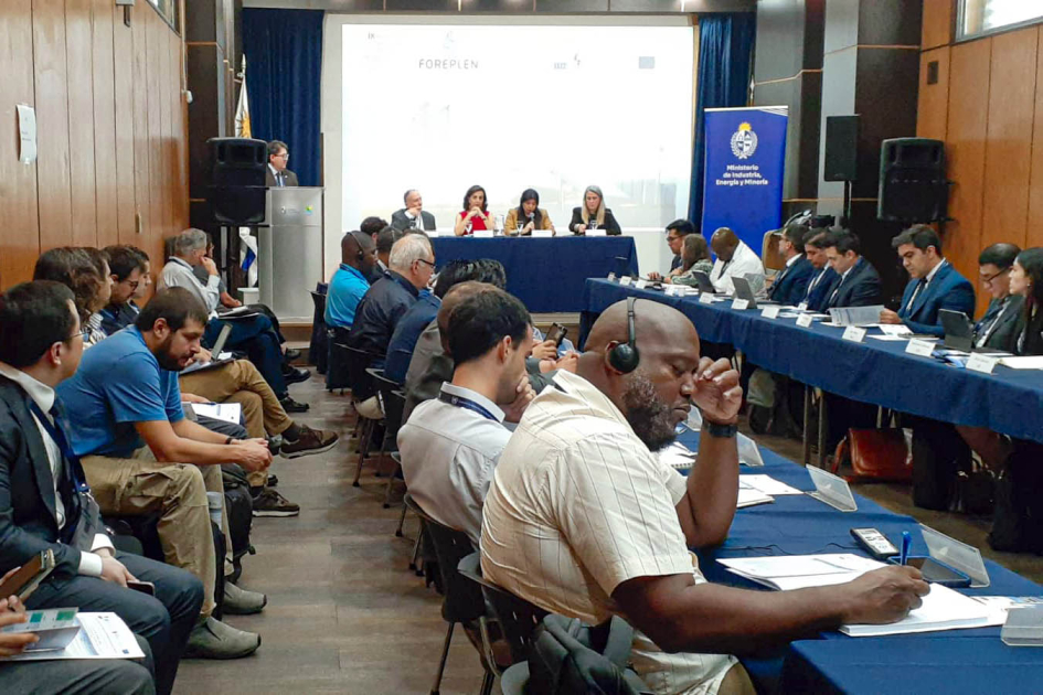 Personas sentadas en mesas enfrentadas y en segunda fila; al fondo, mesa y pantalla