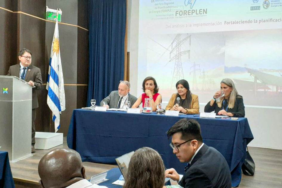 Spinelli habla en mesa de expositores; detrás, pantalla; al lado, hombre de pie en un atril