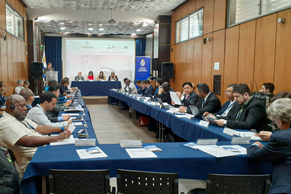 Personas sentadas en forma de "u"; al fondo, pantalla y mesa con personas sentadas