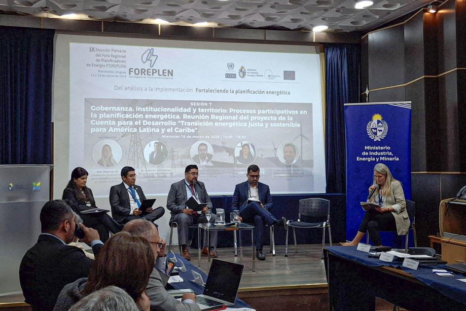 Personas sentadas en un escenario; detrás, pantalla; más cerca, personas en mesas enfrentadas