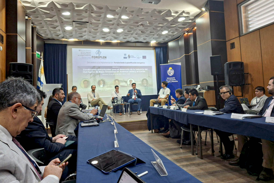 Personas sentadas en un escenario; detrás, pantalla; más cerca, personas en mesas enfrentadas