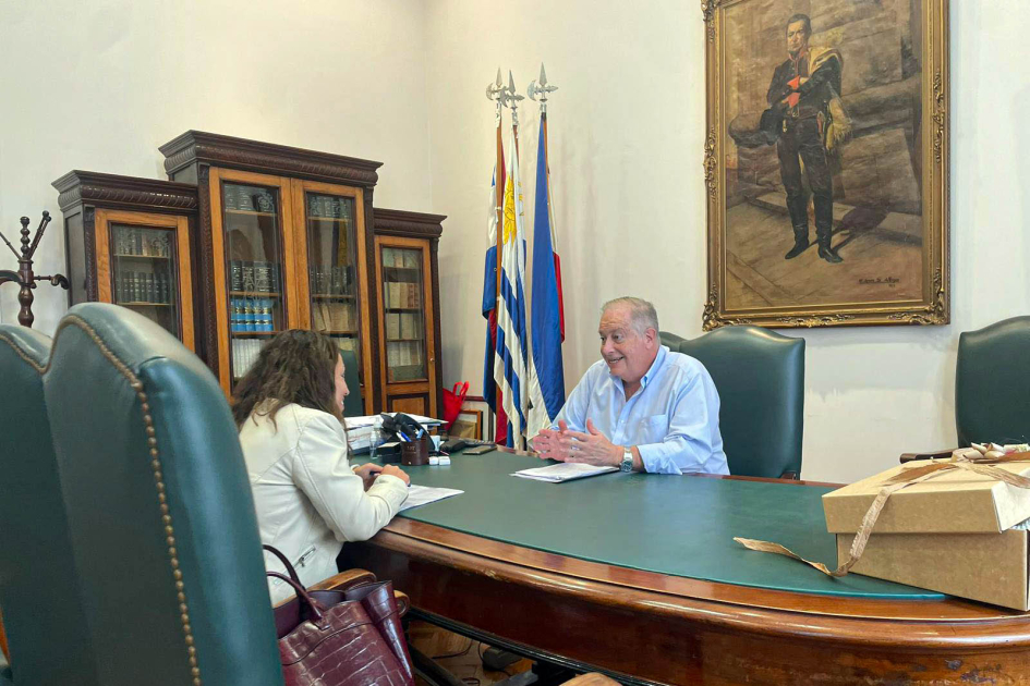 Ezquerra y Cardona dialogan a ambos lados de una mesa; al fondo, cuadro de Artigas y pabellones