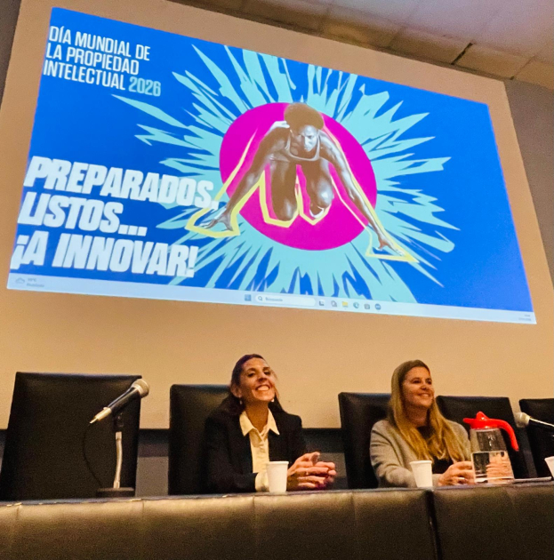 Dos personas sentadas detrás de una mesa con una pantalla de fondo