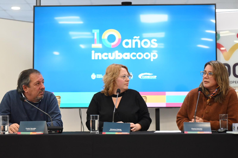 Autoridades en mesa En mesa Ximena Cidrás, Flavia Carreto y Fredy Rondán. Detrás, pantalla con imagen de Incubacoop
