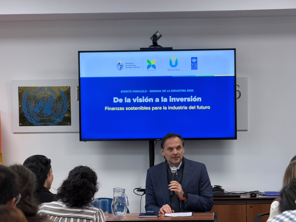 Pettinato habla en una mesa; de espaldas y frente a él, público; encima, pantalla