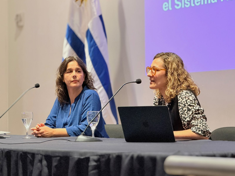 Tomassini y Prieto, sentadas; detrás, bandera uruguaya y pantalla