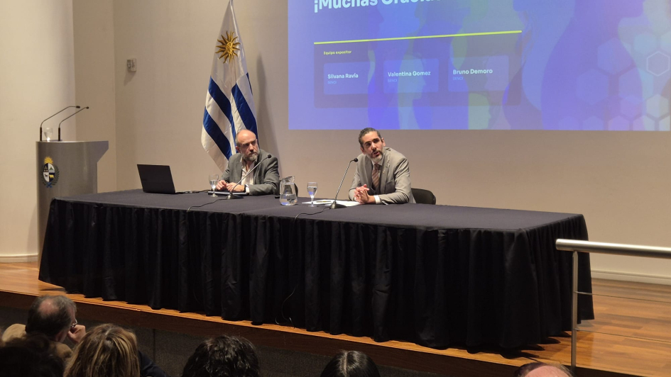 Mesa en que están Díaz y González, sentados; detrás, bandera uruguaya y pantalla