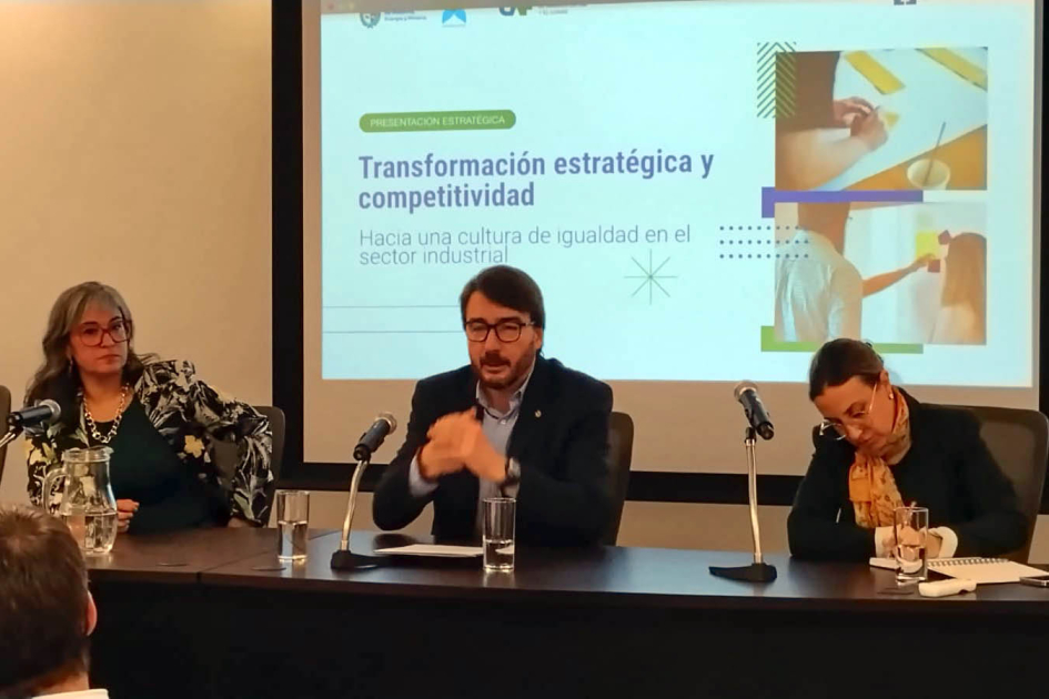 Míguez habla, sentado junto a dos personas; detrás, pantalla