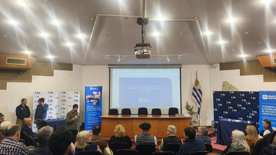 Espacio de intercambio Sala con personas sentadas; al fondo, mesa, pantallay bandera uruguaya