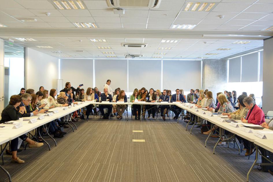 Vista de una gran sala, donde muchas personas están sentadas en mesas que forman un rectángulo