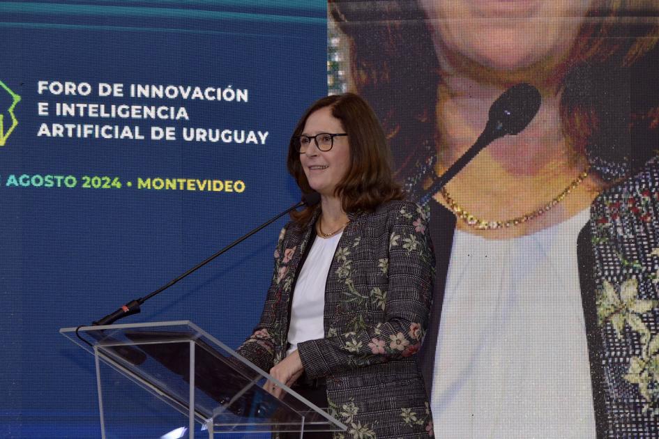 Ministra Elisa Facio Ministra Facio habla desde un atril; detrás, pantalla con parte de su foto y nombre del evento