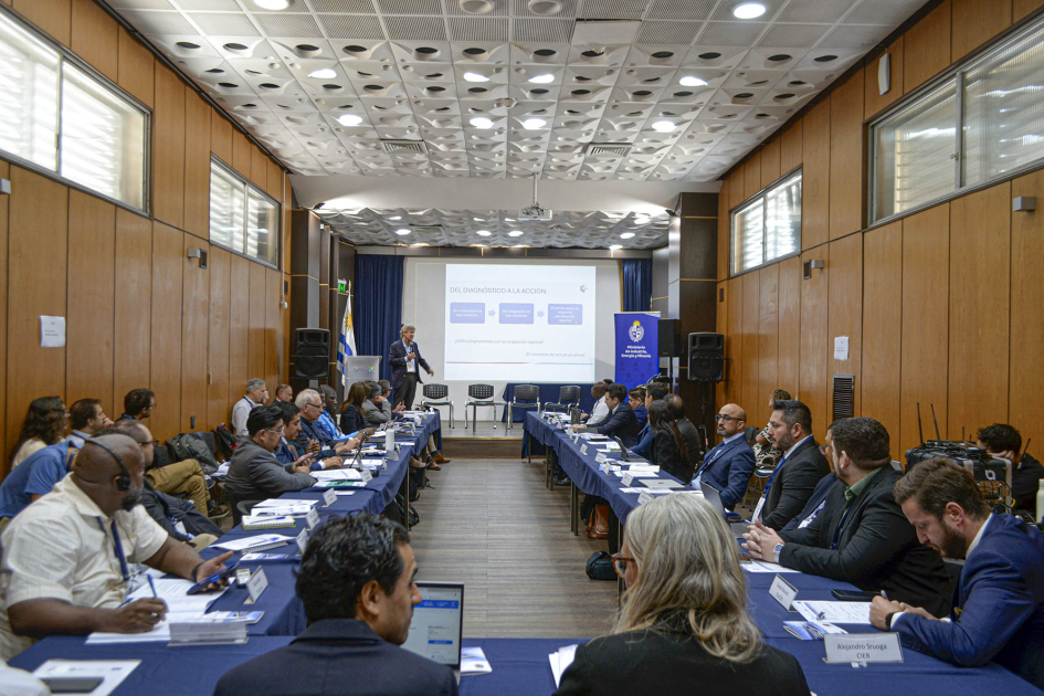 Personas sentadas en mesas en forma de "u"; al fondo, persona de pie y pantalla