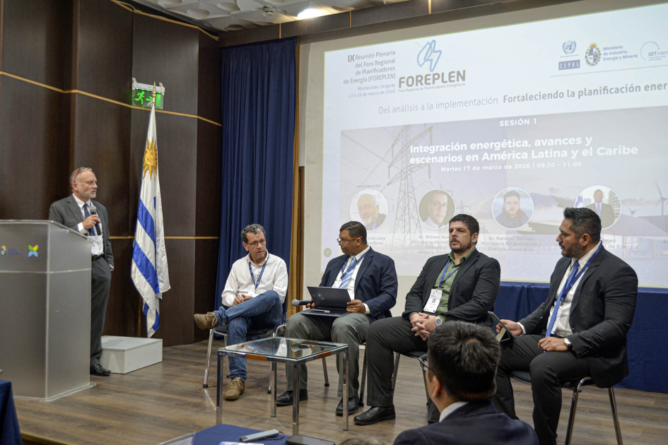 Personas sentadas; detrás, pantalla y bandera uruguaya; de pie, persona habla desde un atril