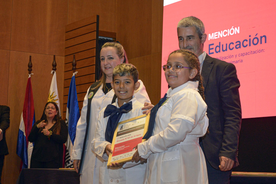 Niñas, niños, docente y autoridad, en entrega de premio; detrás, pantalla