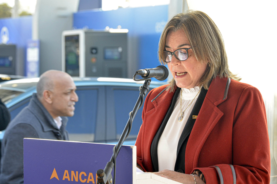 Sandra Saffores habla a micrófeno, detrás se ve un hombre y un auto.