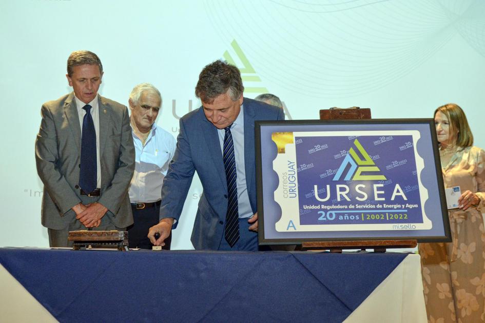 Paganini matasella durante la presentación del sello conmemorativo del Correo Uruguayo Pagani matasella sobre una mesa que tiene el sello conmemorativo de URSEA; a su lado está Navarrine