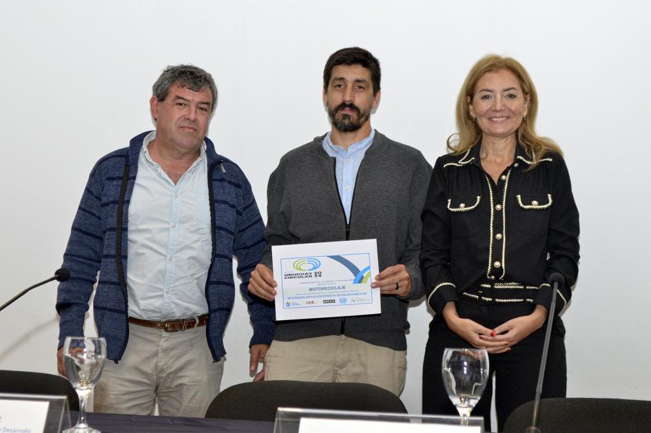 Mención especial de la categoría Cooperativas, Asociaciones y Organismos Públicos: Motoreciclaje Directora Natalia Jul, de pie, junto a dos hombres; uno sostiene un diploma