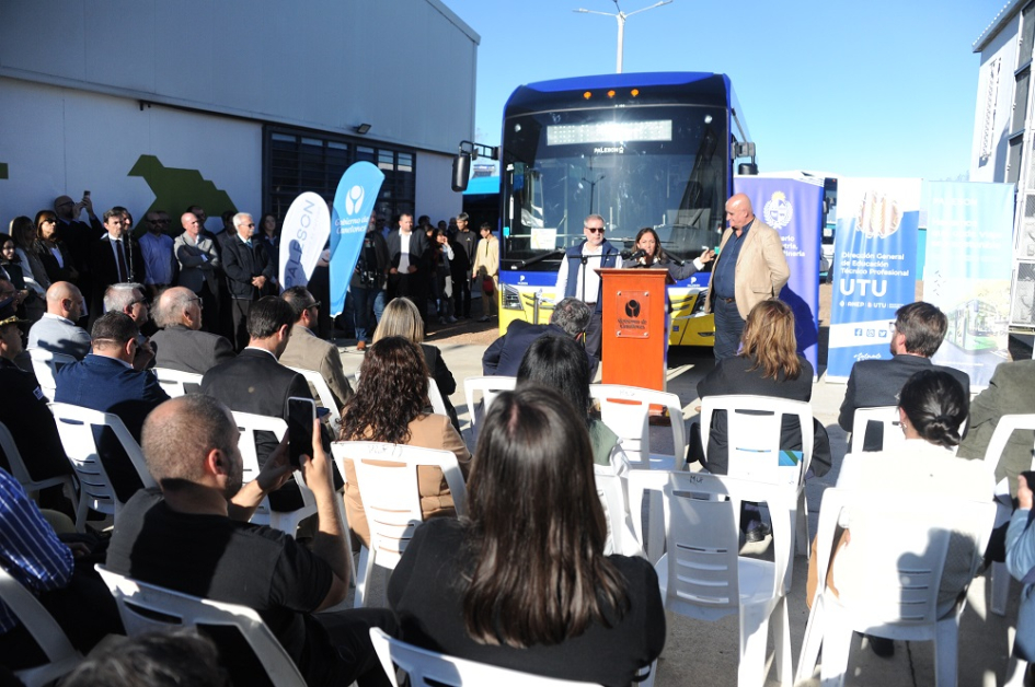 Público sentado. Al fondo, Cardona habla desde atril. Detrás, un ómnibus eléctrico 