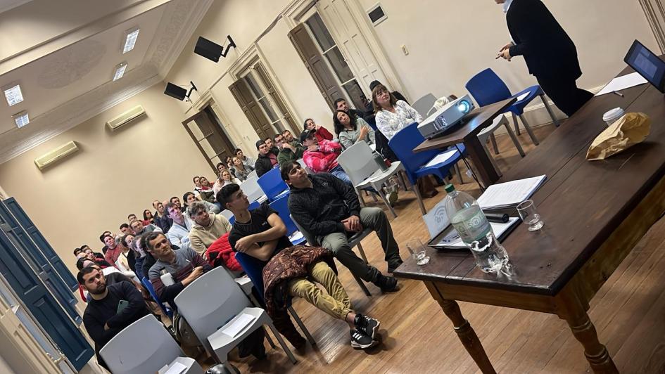 Participantes del curso MPersonas sentadas en un salón; más cerca, un docente de pie y una mesa