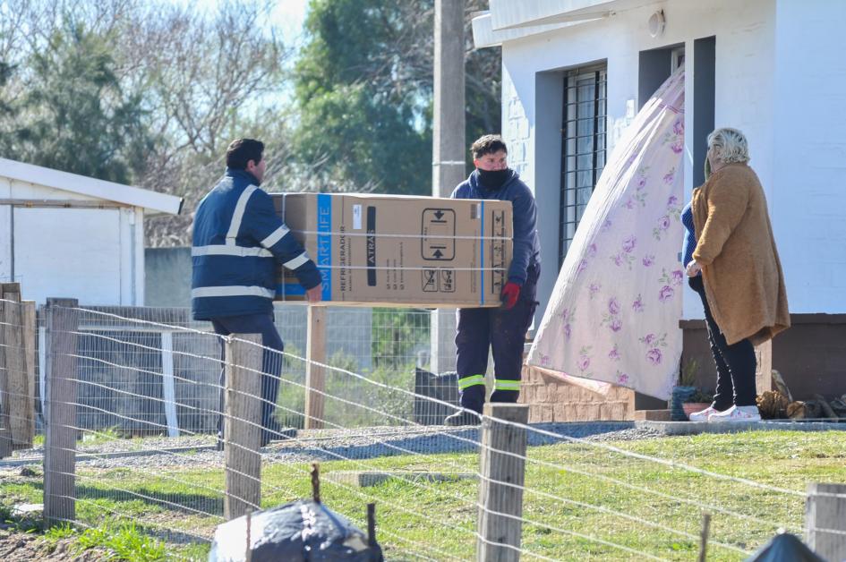 Entrega de una heladera en el hogar de una beneficiaria Personas cargando una heladera hacia la casa de una beneficiaria