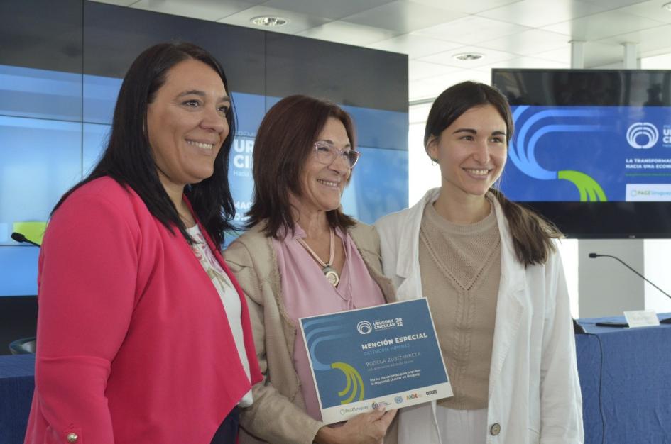 Mención en categoría Mipymes: Bodega Zubizarreta Sánchez junto a dos mujeres; una sostiene un diploma; detrás, dos pantallas