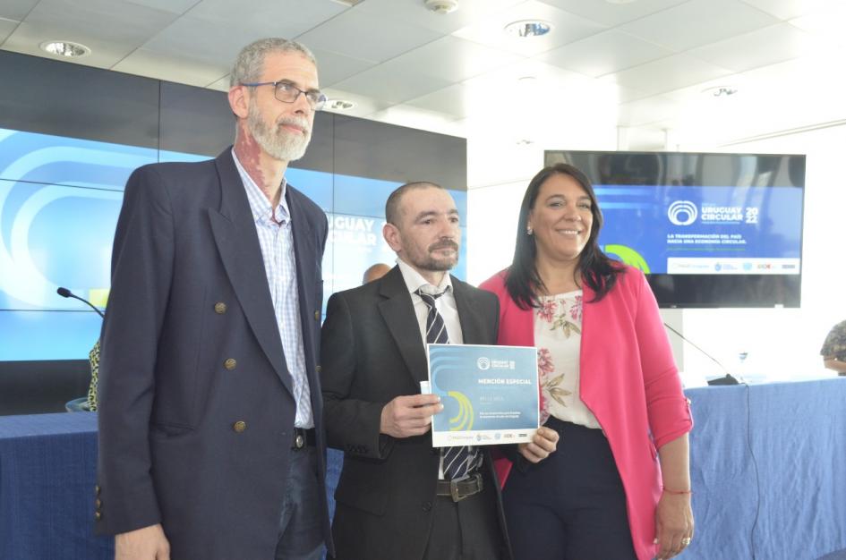 Sánchez junto a dos hombres; uno sostiene el diploma. Detrás se ven dos pantallas