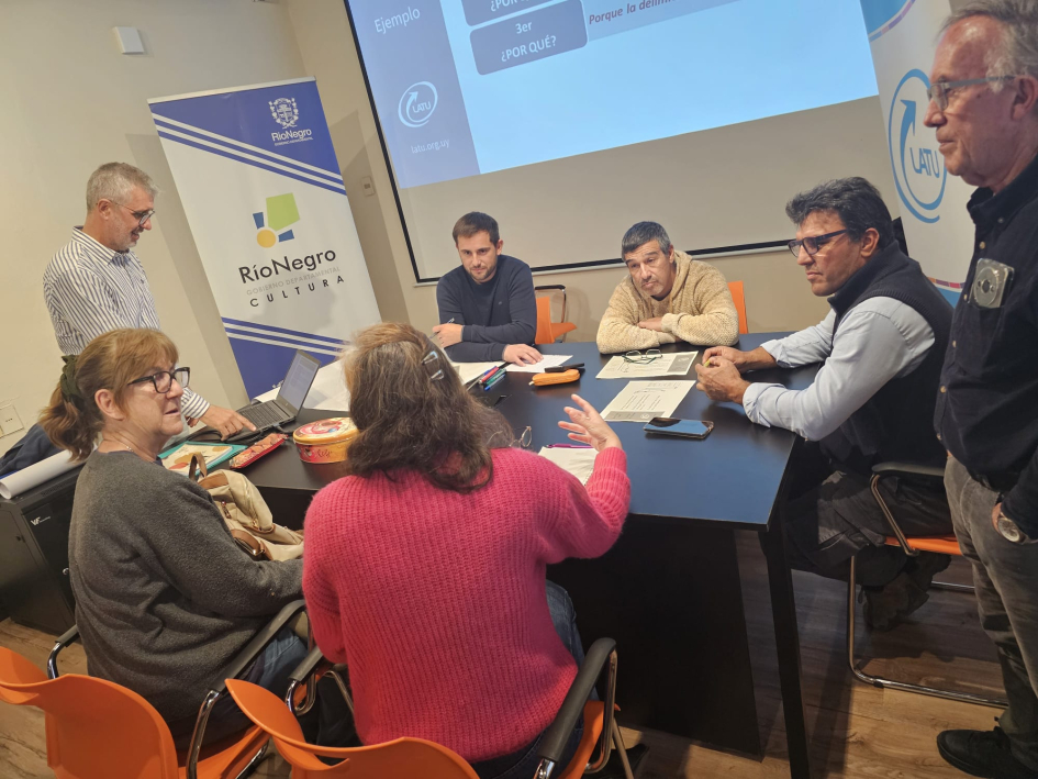 Intercambio de experiencias Personas sentadas alrededor de una mesa; al lado, personas de pie, pantalla y banner institucional
