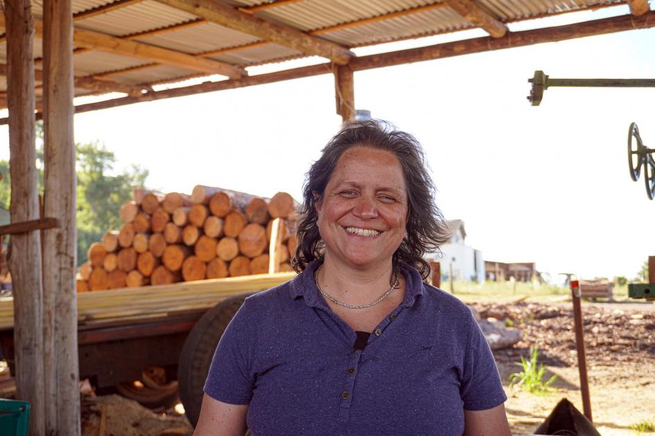 Una mujer sonríe a la cámara; detrás de ella hay un camión con madera