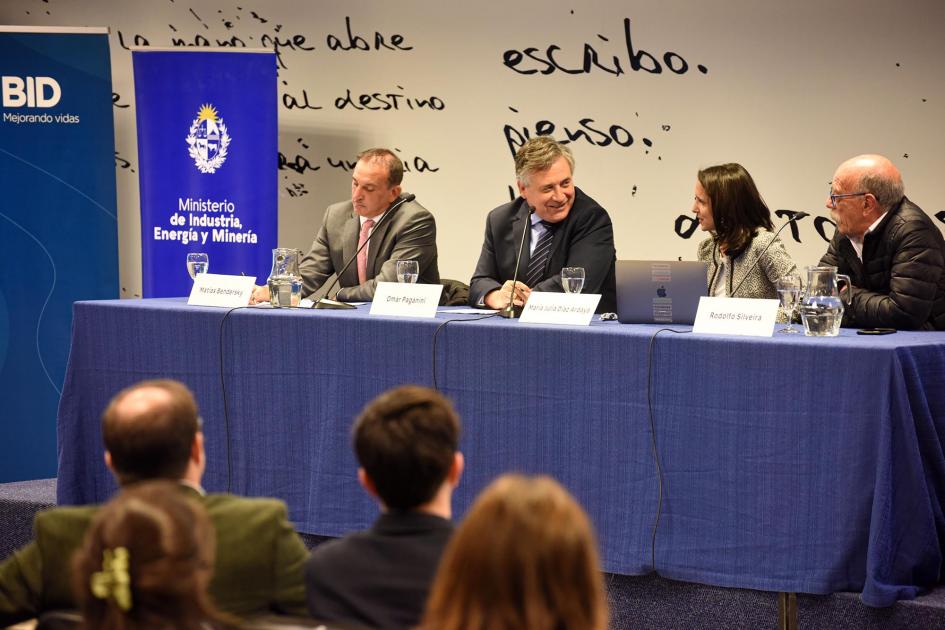 Mesa de autoridades; Paganini sonríe; delante se ve el público; al lado, banners