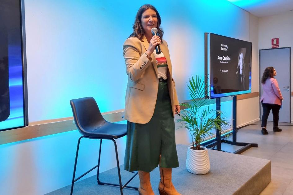 Ana Castillo, de BID LAB Una mujer habla sobre un escenario; a sus lados, una silla y pantallas; a lo lejos se ve otra muje