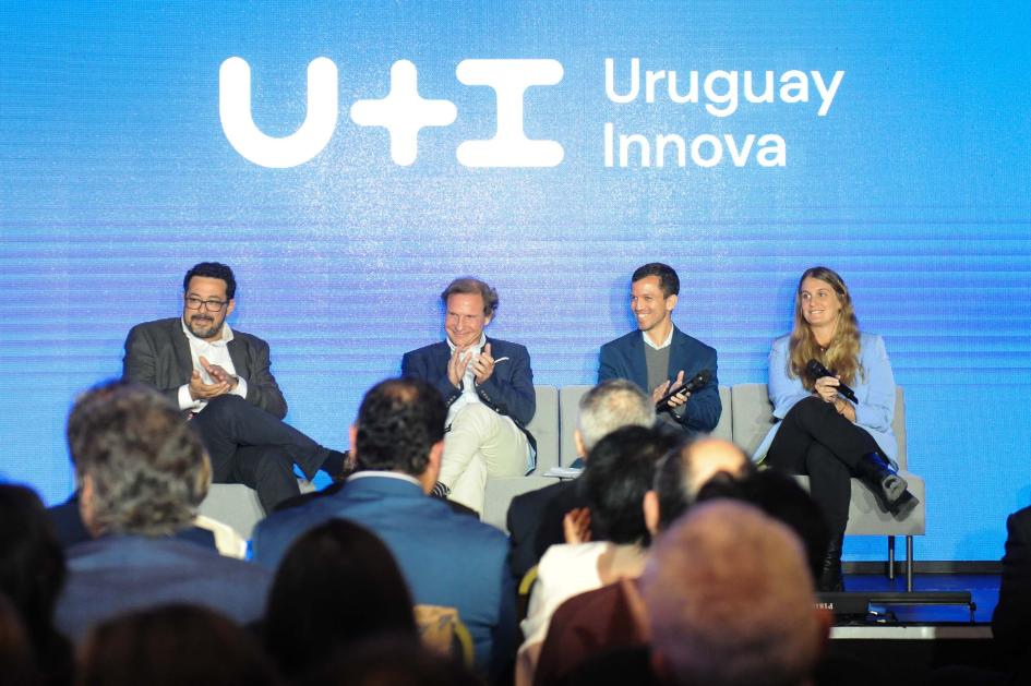 Público de espalda; al fondo, panel del evento; detrás, pantalla azul con logo del programa