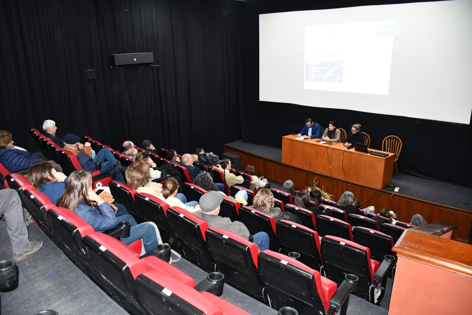 Al fondo, mesa de autoridades y pantalla más cerca, personas sentadas en auditorio