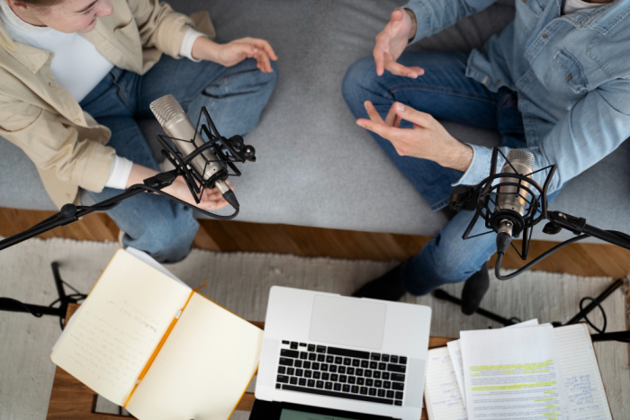 Dos personas sentadas en el piso hablan por micrófonos, entre una computadora y apuntes