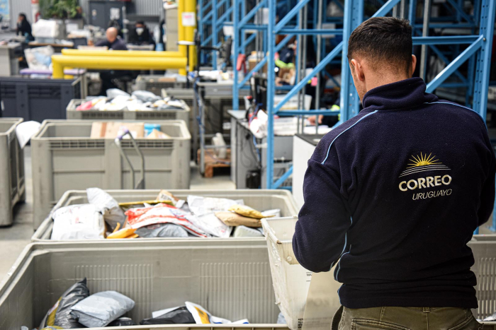 Persona con una campera del Correo Uruguayo, de espaldas, trabaja con cajas con objetos a su lado