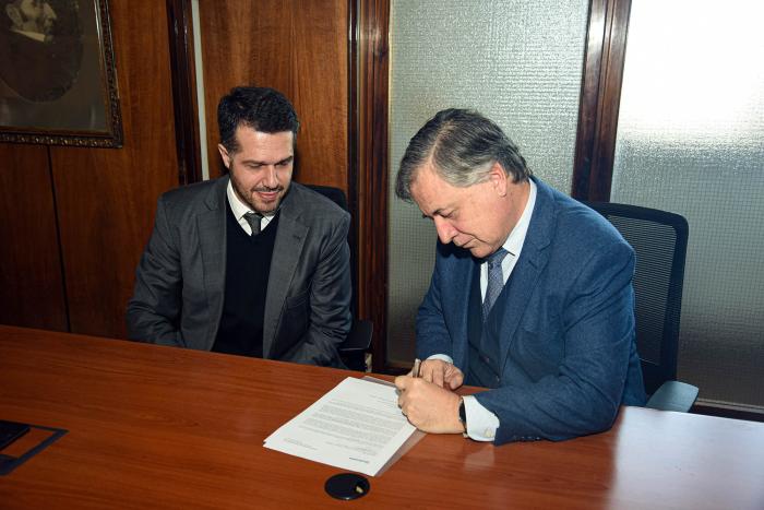 Firma del documento Paganini firma un documento, sobre una mesa, junto a otro hombre
