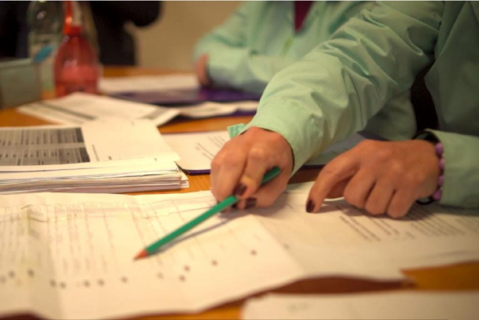 Manos de persona, con un lápiz, sobre documentos; detrás, se ven manos de otras personas