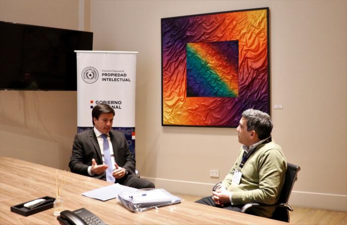 Dos hombres están reunidos en una mesa. Detrás hay un cuadro de colores