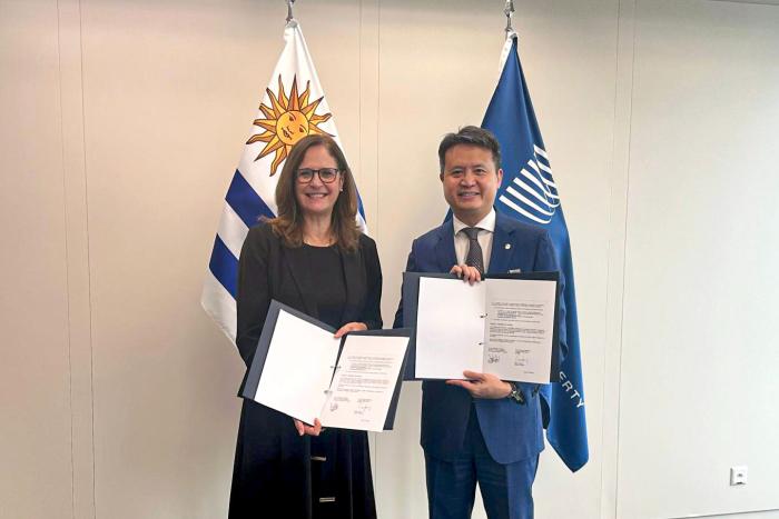 Facio y Tang, de pie, sostienen los documentos firmados; detrás, banderas de Uruguay y de la OMPI