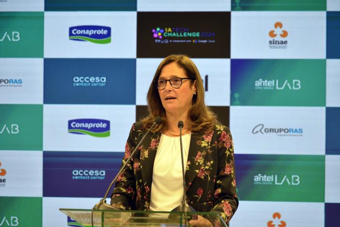 Ministra Facio habla con un micrófono; detrás, banner de instituciones