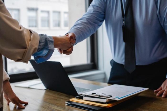 Dos personas se dan la mano. Entre ellos hay una mesa y una laptop