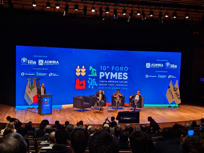 Panel del 10.° Foro Pymes Italia-América Latina Personas en un escenario; detrás, pantalla; más cerca y de espaldas, público