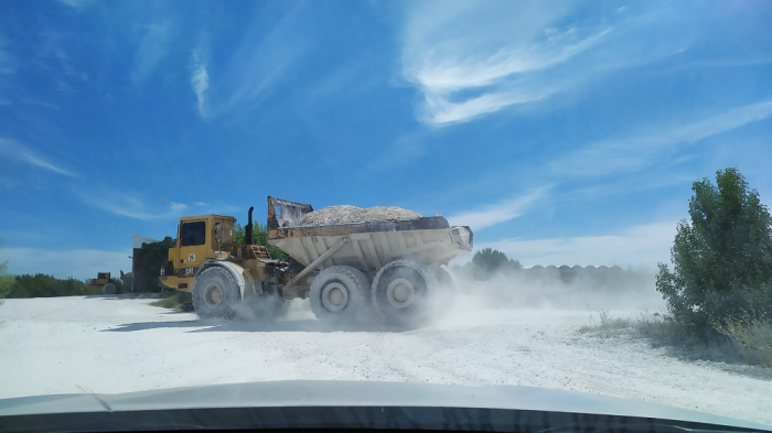 En paisaje con cielo azul y suelo arenoso con algunos arbustos, un camión cargado de rocas