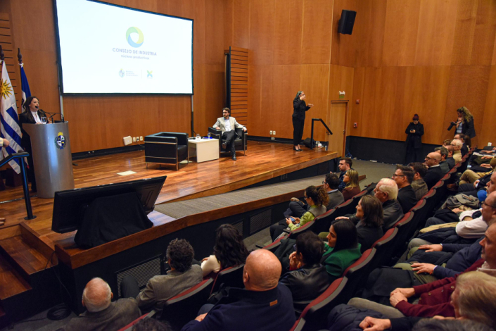 En estrado, Fernada Cardona en atril y director Adrián Míguez sentado. Se ve público en auditorio.